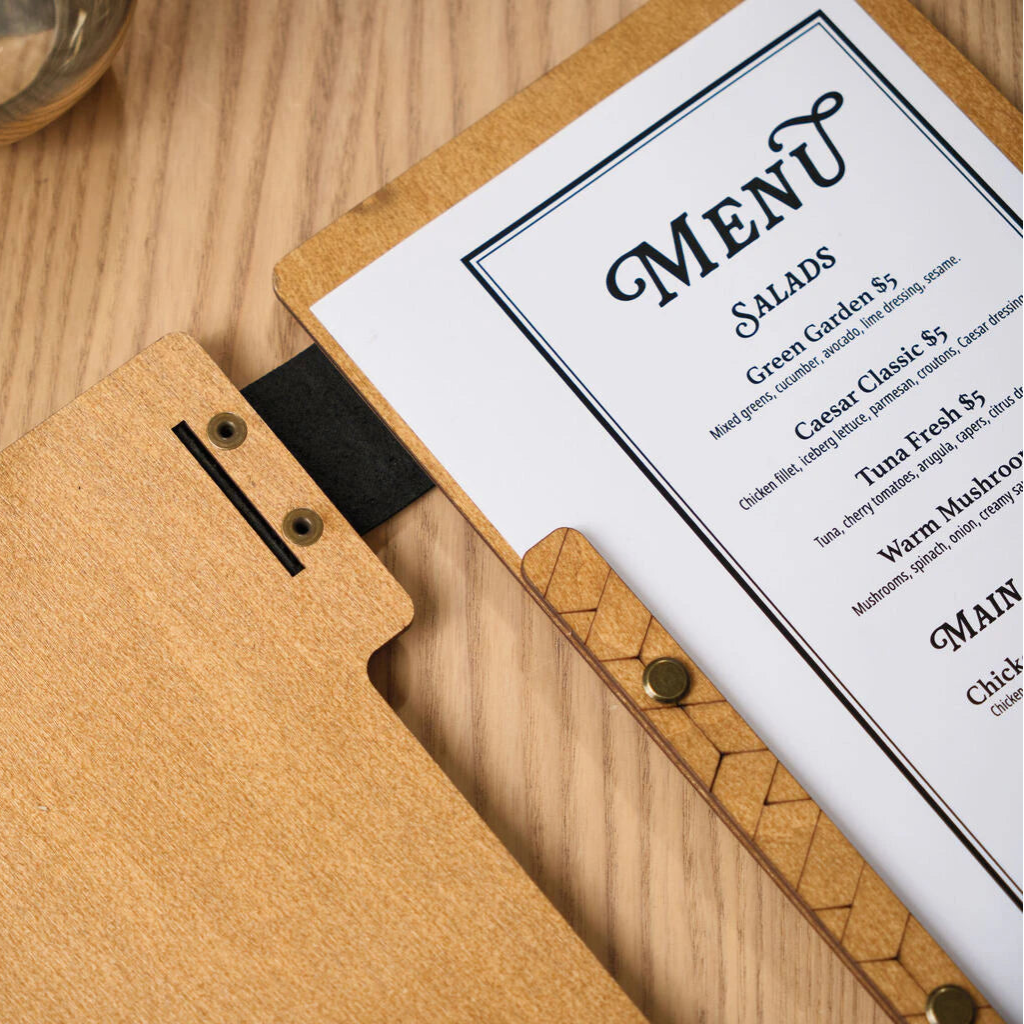 A close-up view of an open oak-colored wooden menu folder, showing the internal binding strip with an engraved geometric pattern and brass screws that hold the printed menu page in place.