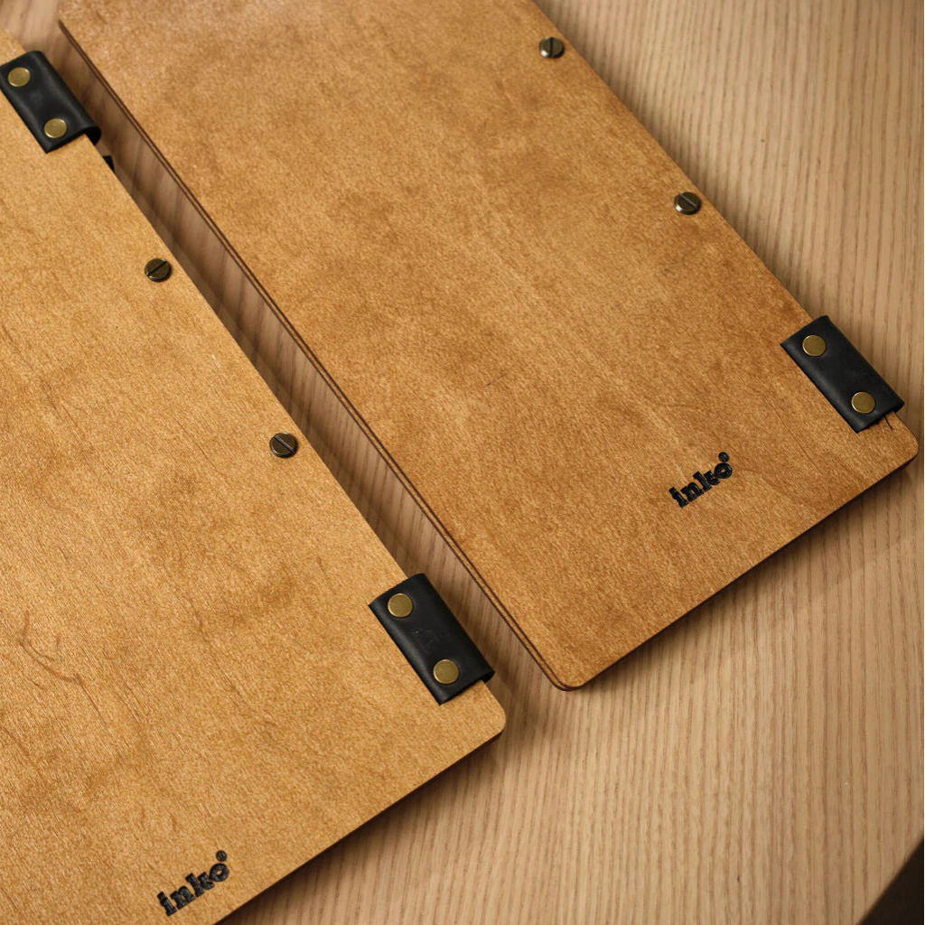 A view of the back of two oak-colored wooden menu folders, showing the smooth wood surface, black leather corner accents with brass rivets, and the small black "inko" logo at the bottom.