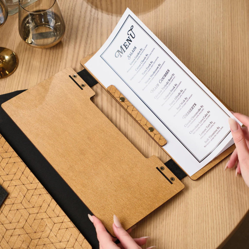 A person's hands holding an open oak-colored menu folder, revealing the clean layout of the printed menu and the textured geometric engraving on the folder's inner binding strip.