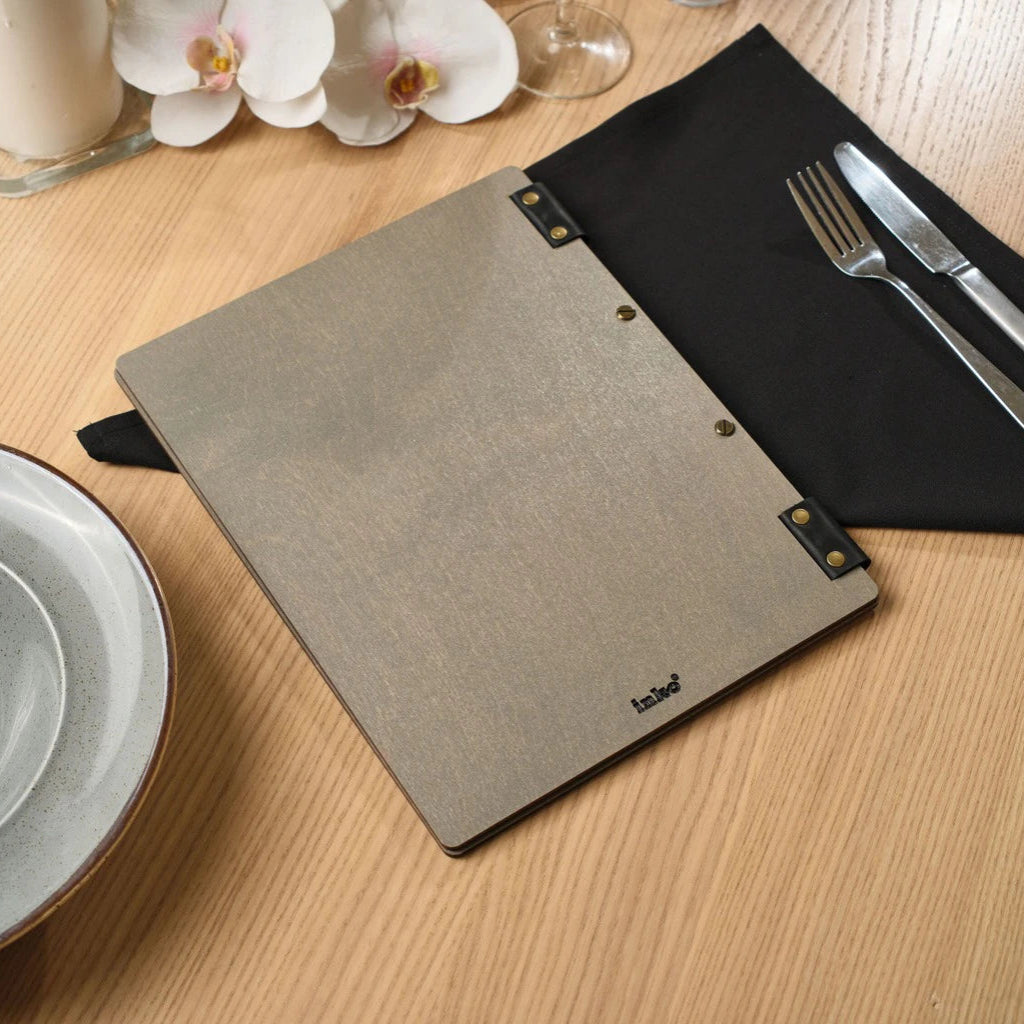 A closed grey wooden menu folder with a subtle grain texture, featuring a black "inko" logo in the bottom corner and leather-and-brass hardware, displayed on a light wooden table with restaurant settings.
