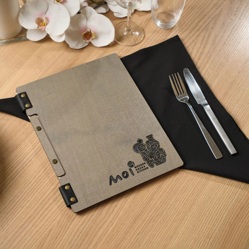 Custom grey wooden menu folder for "Moi Modern Hakka Kitchen". This handcrafted wood menu cover features a unique black leather strap binding, laser-engraved branding, and bronze hardware on a modern restaurant table setting