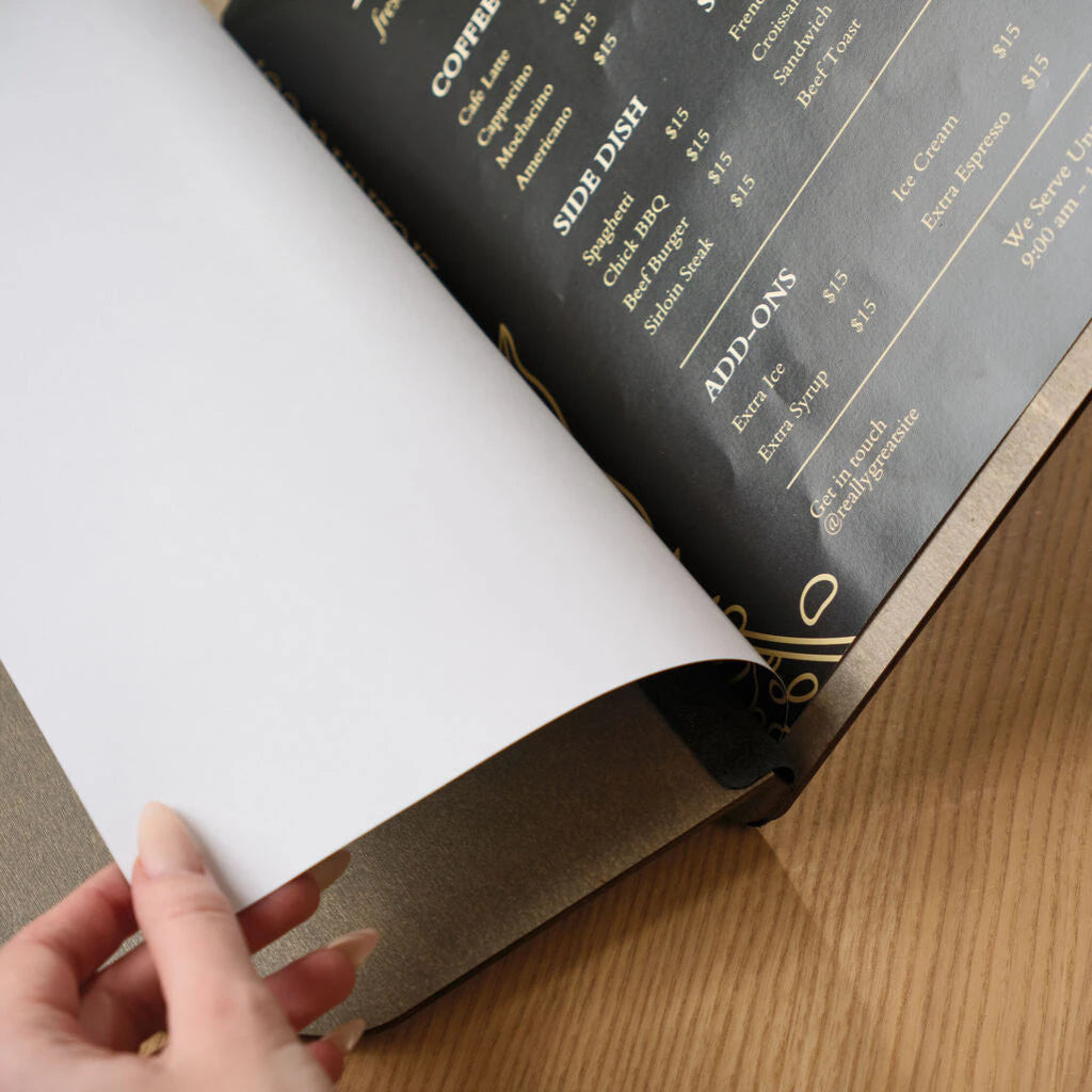 Detail view of a hand lifting a white page within a custom wooden menu holder, showing the flexible binding and premium paper quality