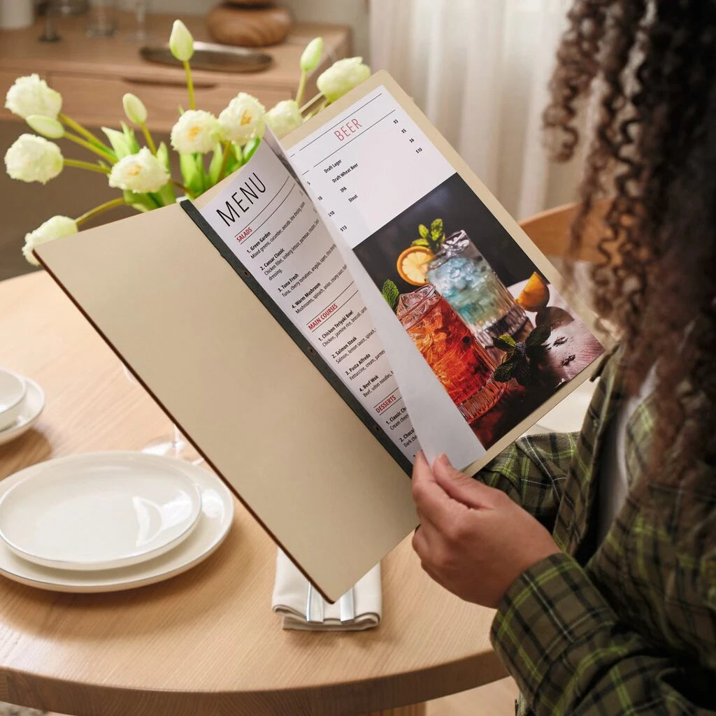 Woman reading open wooden menu holder with cocktail photos at restaurant table with white tulips