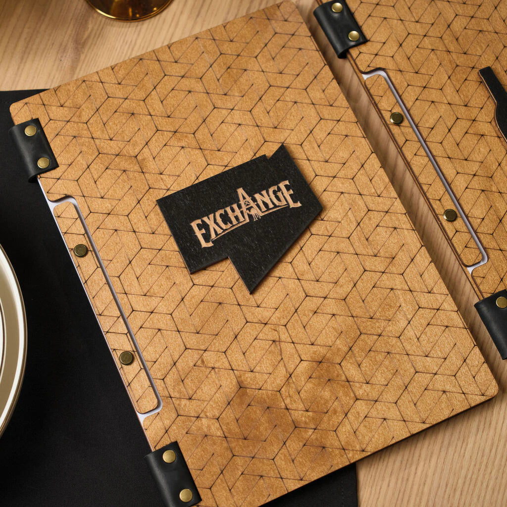 Close-up of a wooden menu cover with laser-engraved intricate geometric patterns and a raised wooden "EXCHANGE" logo with black leather fasteners