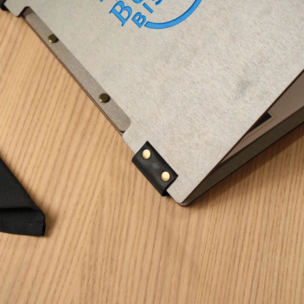 Close-up view of the corner of a grey wooden menu folder, focusing on the fine wood texture and the black leather strap secured with gold-colored metal studs.