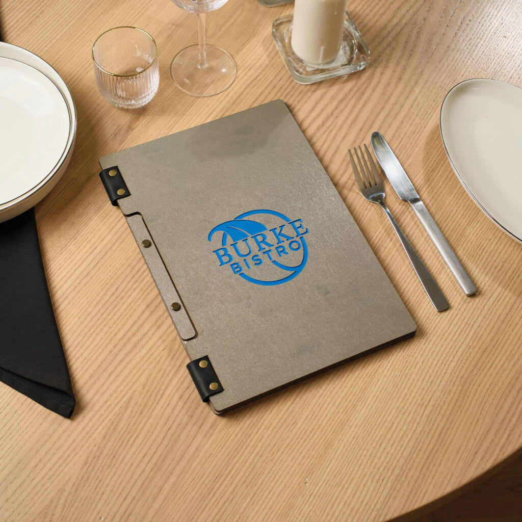 A closed grey ash wood menu folder on a restaurant table, featuring a blue "BURKE BISTRO" logo. The folder is bound with black leather hinges and gold-toned brass rivets.