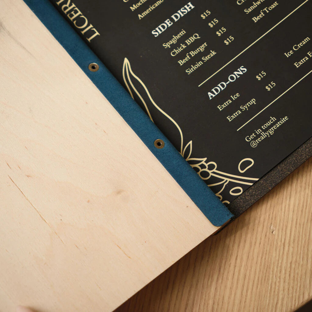 Macro detail of the blue Jeans-colored spine on a wooden menu board, showing the integrated brass eyelets and the smooth transition to the Ivory wood cover