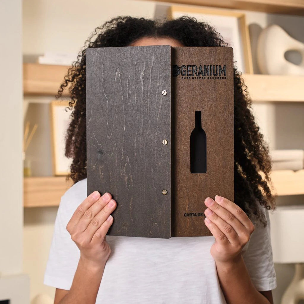 A person holding the menu folder upright in front of their face, showcasing both the solid back cover and the decorative front cover with the bottle-shaped window.