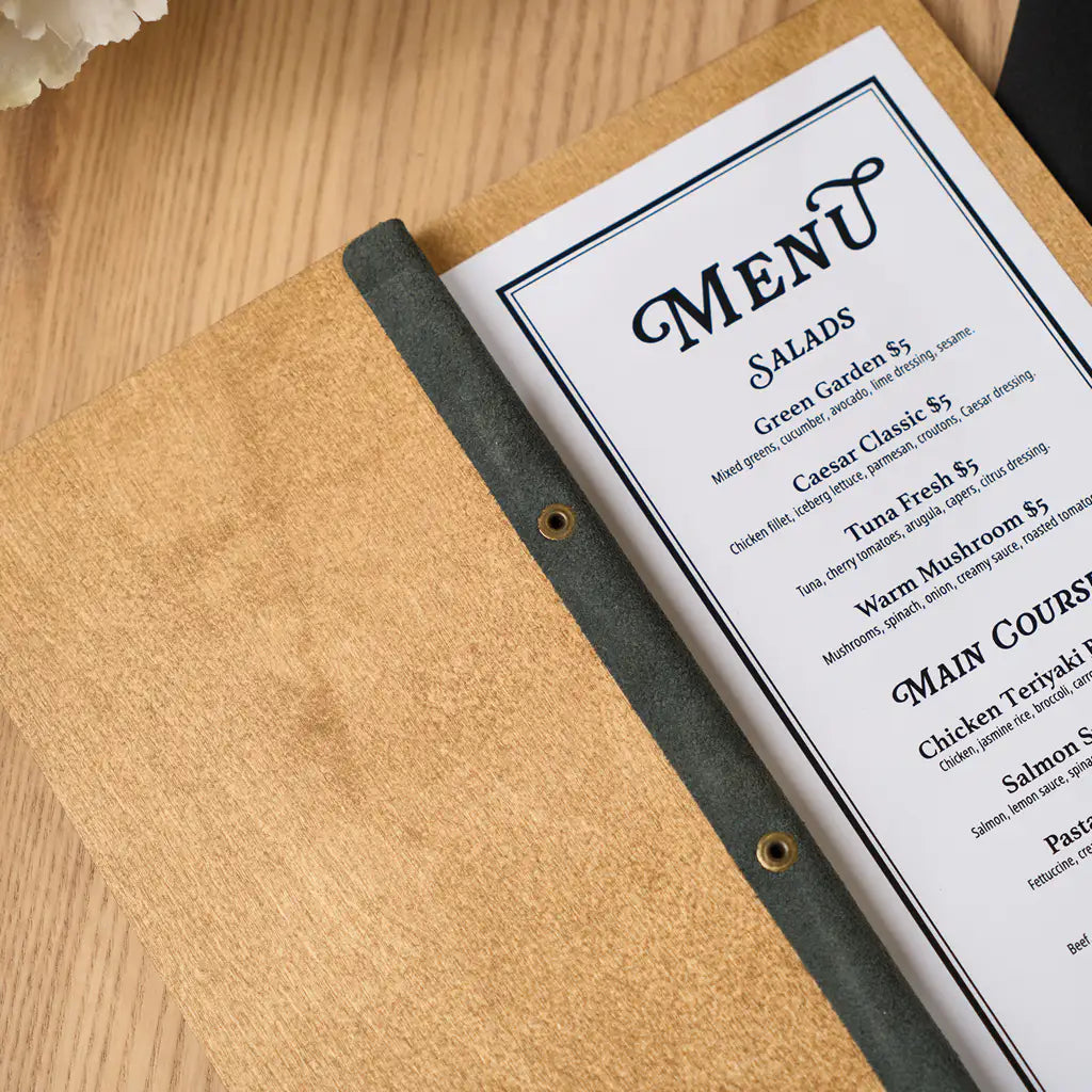 An open oak-colored wooden menu folder resting on a black napkin, displaying a printed menu insert with sections for Salads, Main Courses, and Desserts.