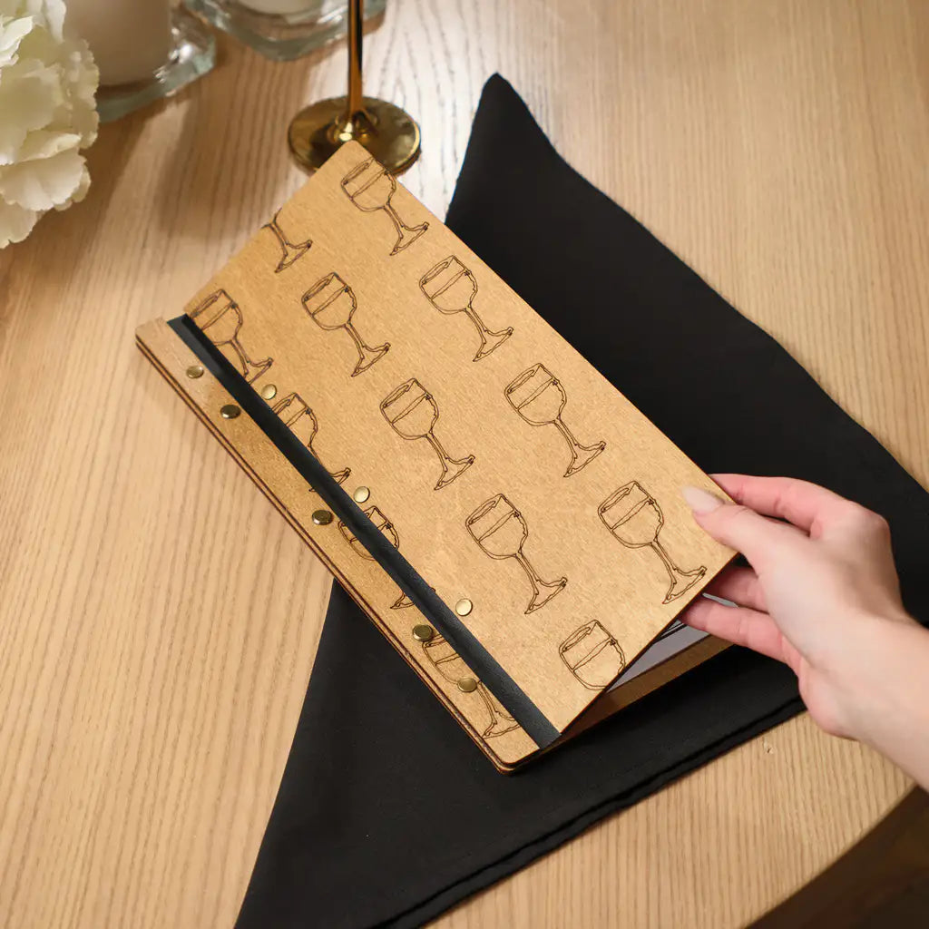 A person’s hand lifting the front cover of a light-colored wooden menu folder, revealing an engraved pattern of wine glasses on the oak surface.