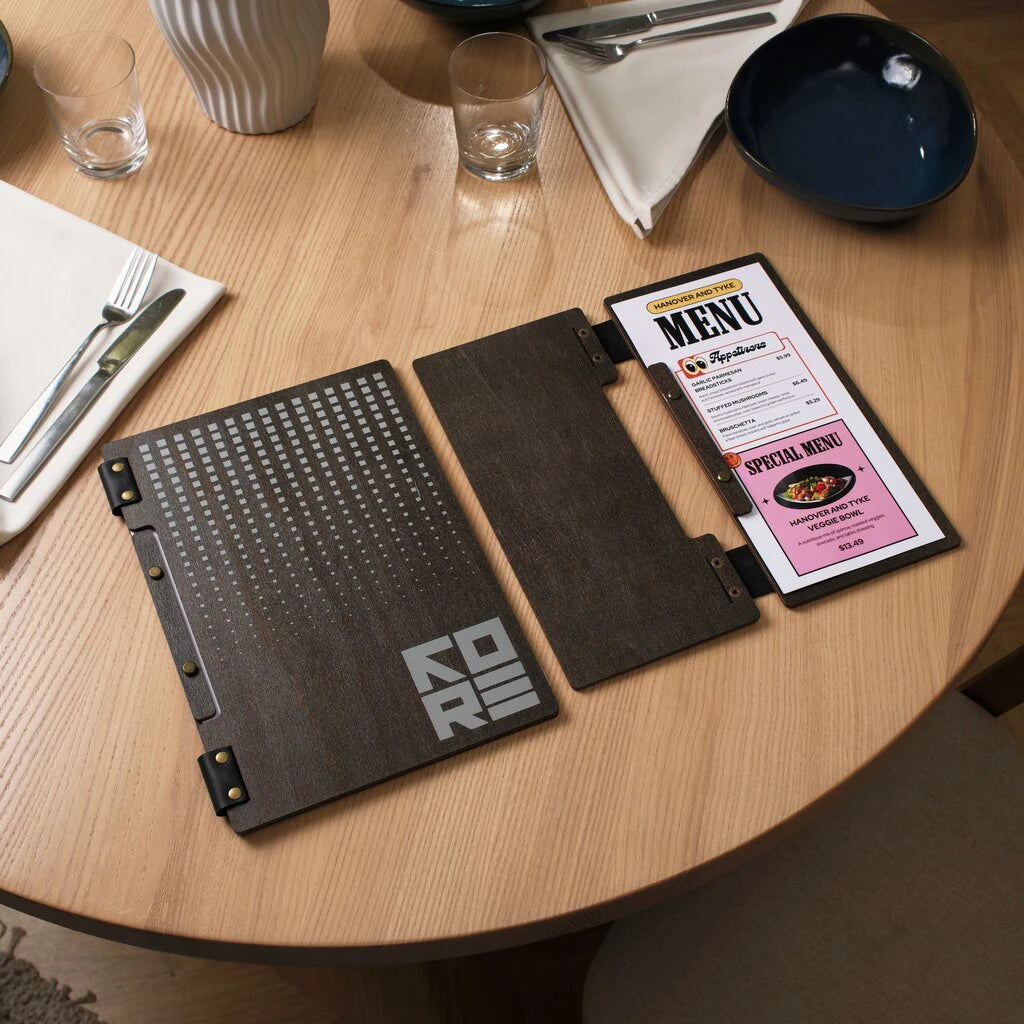 Lifestyle shot focusing on a narrow wooden drink or special menu holder with a flexible leather spine. The image shows the contrast between a standard-size wooden menu cover and a compact, elongated version. Both folders feature laser-engraved geometric patterns and branding, ideal for organized table service in modern restaurants