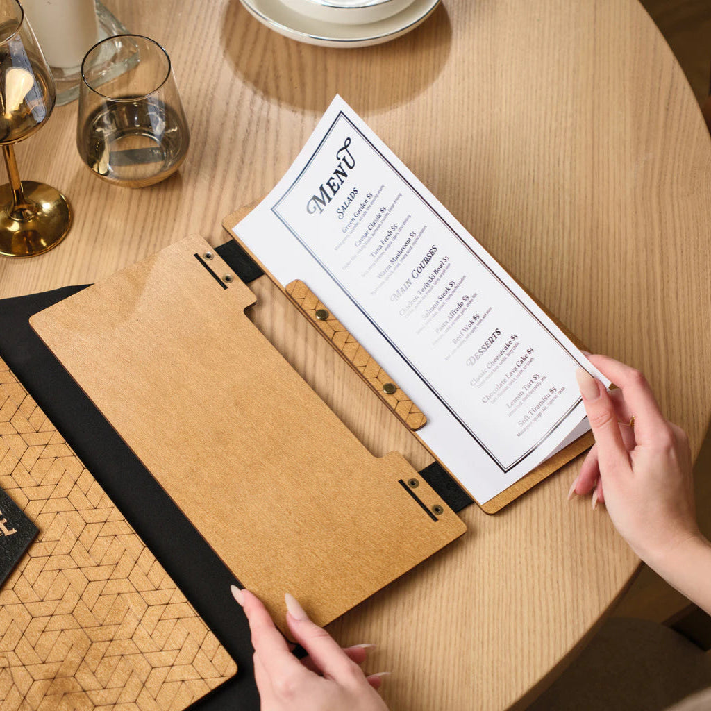 A person’s hands holding open an oak wooden menu folder to reveal a printed menu list. The folder uses black leather hinges and brass hardware to secure the pages.