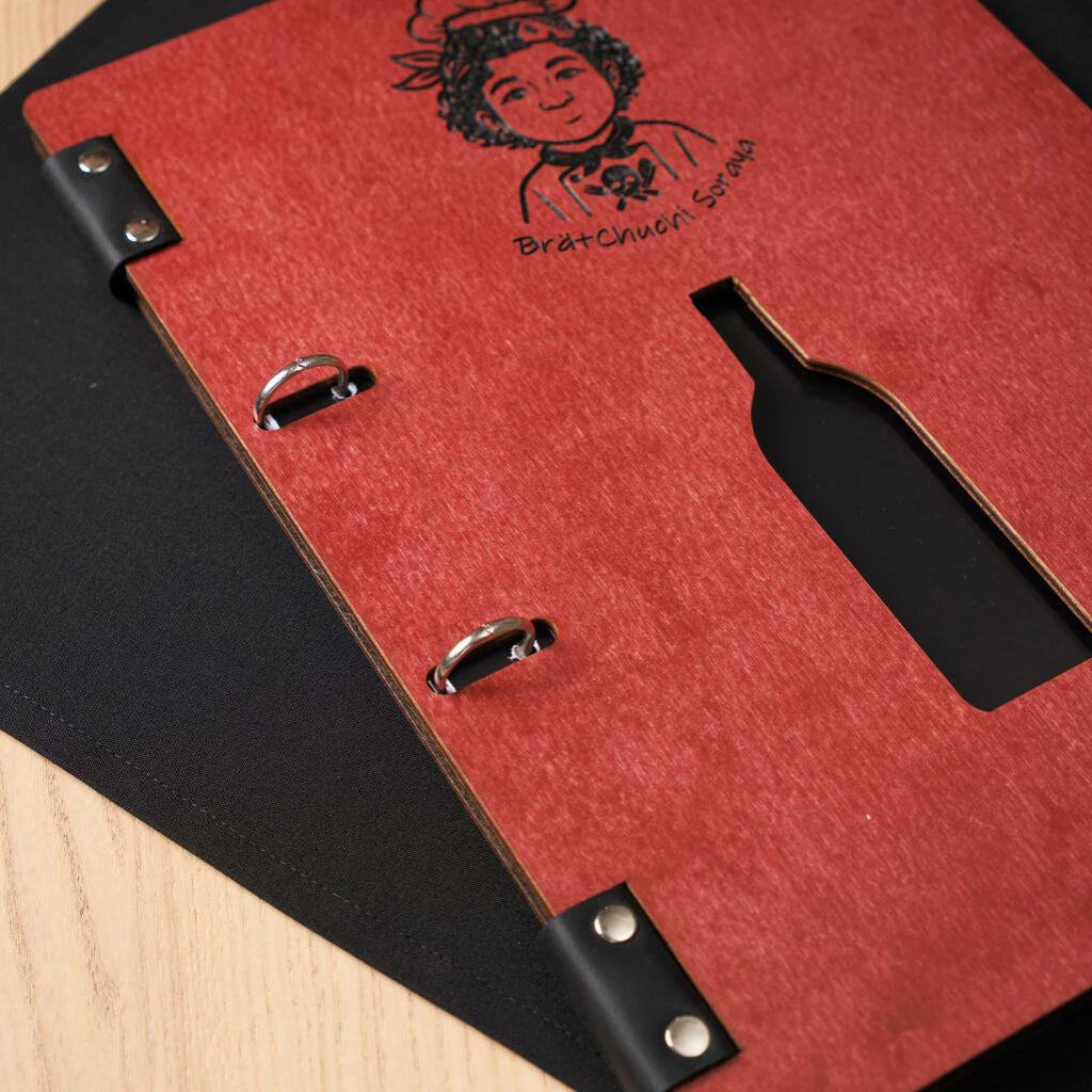 Close-up of the front cover of a chili-red wooden menu, focusing on the silver ring binder clips and the clean, laser-cut silhouette of a wine bottle.
