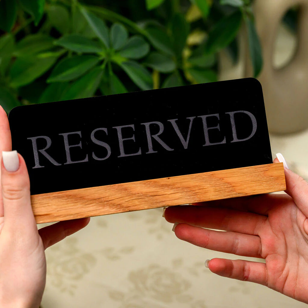 Hand holding a premium "Reserved" table tent sign featuring a black matte finish and oak wood holder on a floral tablecloth background
