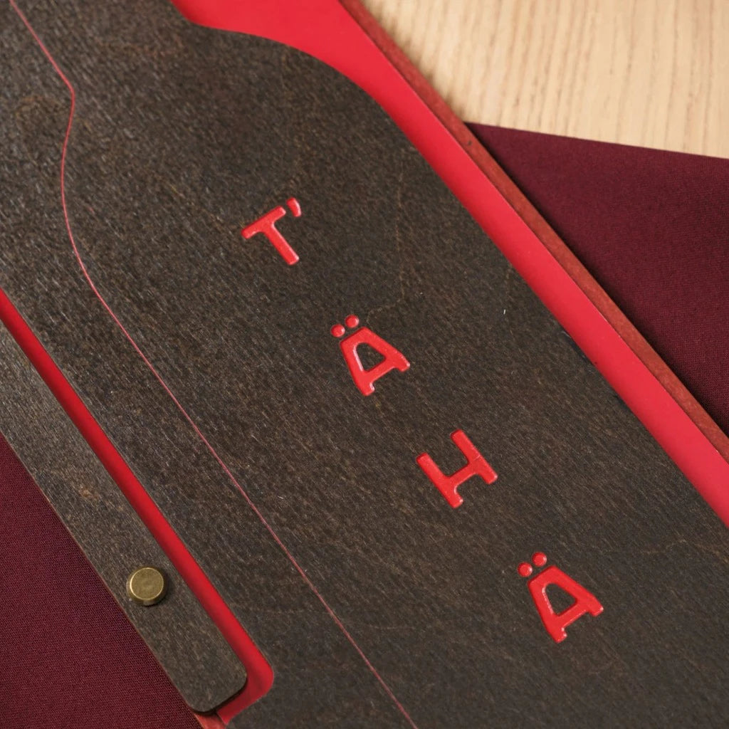 Extreme close-up of a wooden menu folder with vibrant red color engraving and a wine bottle shaped cutout. Shows the intricate wood grain texture, precision laser cutting, and custom branding for high-end restaurants
