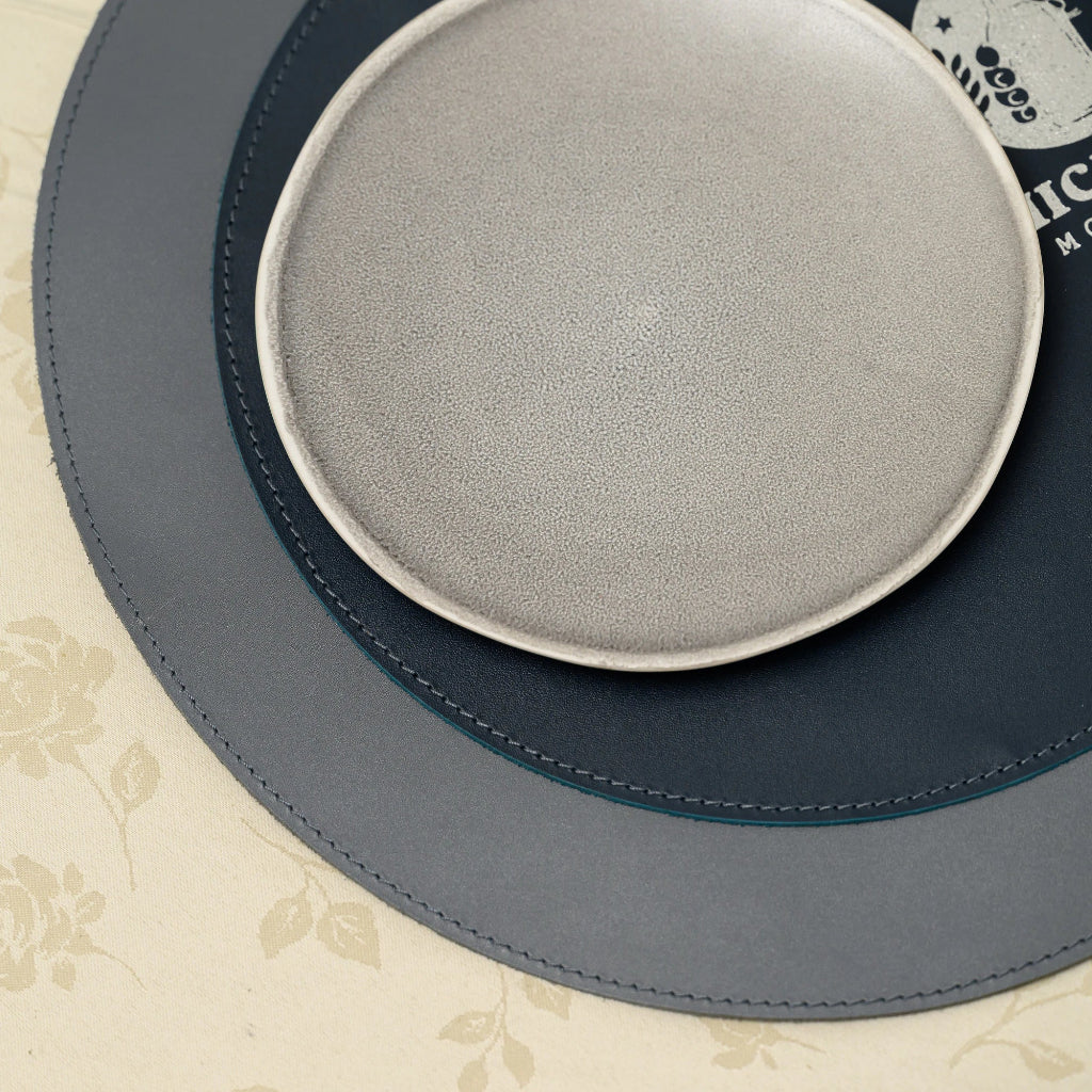 A top-down close-up of a layered leather placemat with a circular design. It features a smooth outer ring joined to a textured inner section by a clean row of perimeter stitching. A small, light-colored ceramic plate rests on the surface, which is set on a floral-patterned tablecloth.