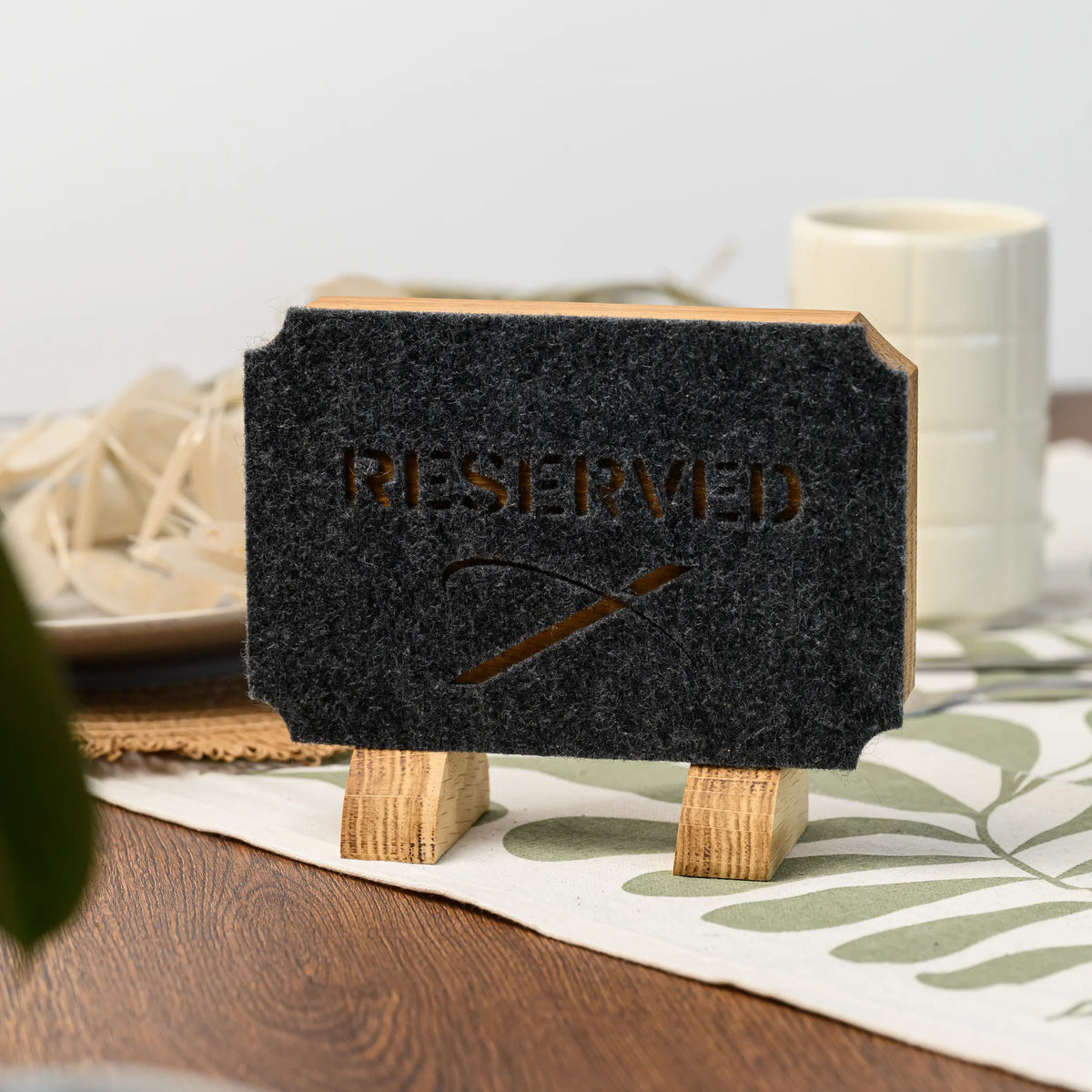 Дубова табличка «Reserved» з прорізаним фетром на столі поруч із піалою, скошені кути фетру повторюють форму дубової основи