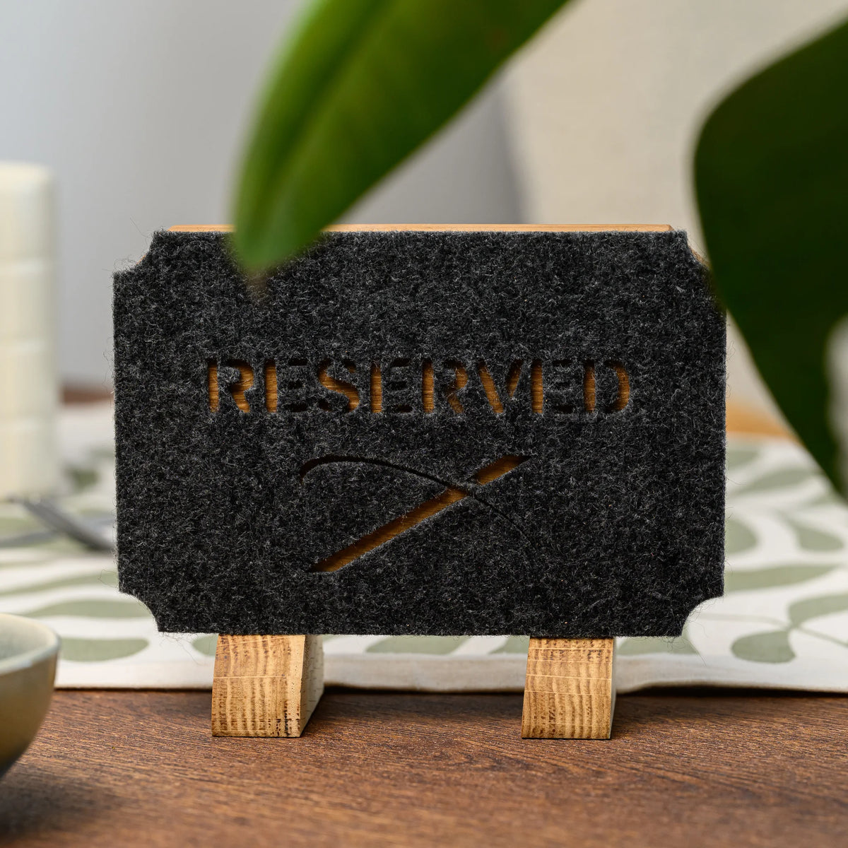 Дубова табличка «Reserved» з прорізаним фетром на двох дубових опорах, вигляд спереду на столі за листям рослини на передньому плані