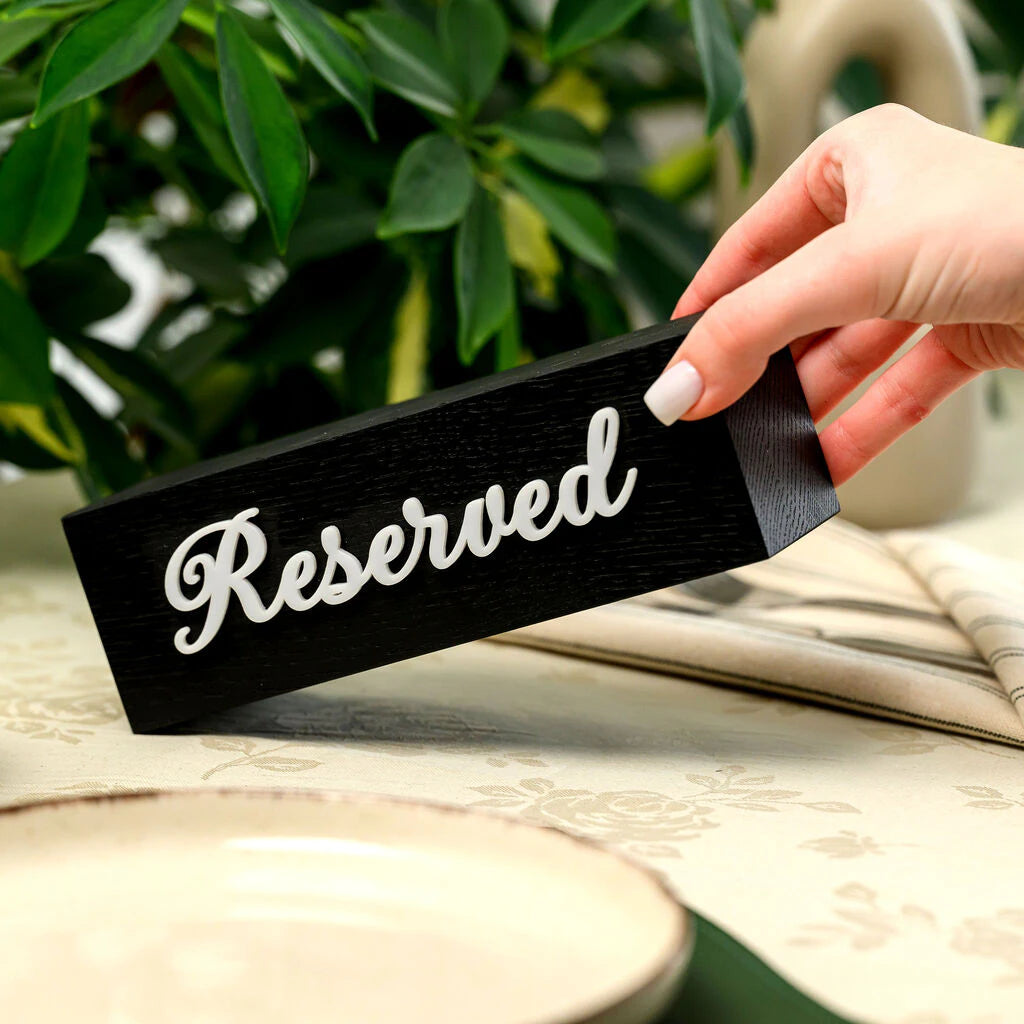 A horizontal "Reserved" table sign held in two hands to demonstrate its scale. The sign is made of high-quality black-stained wood with a prominent wood grain texture and features elegant 3D white acrylic script lettering. It is displayed in a restaurant setting with a neutral tablecloth and greenery in the background