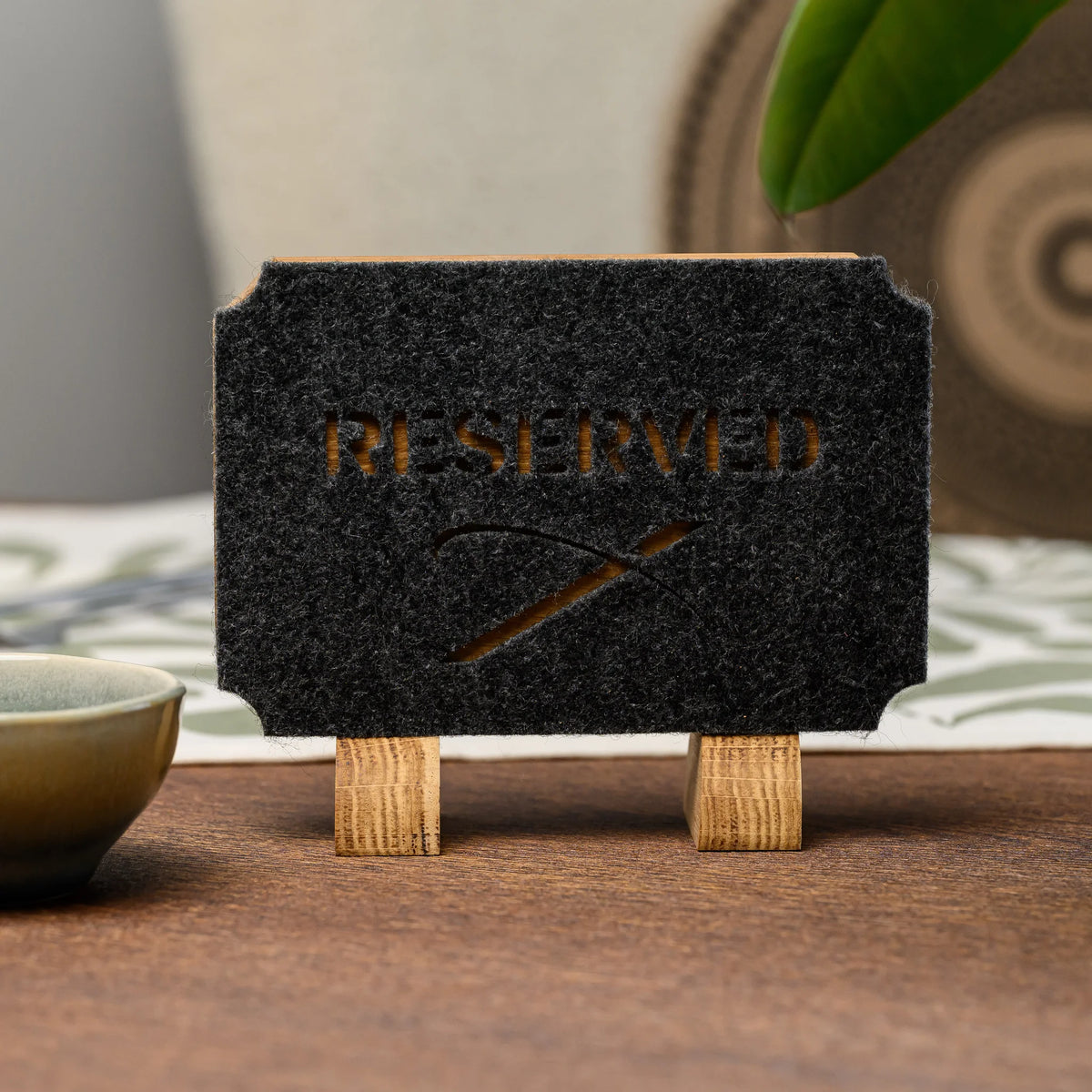 Дубова табличка «Reserved» з прорізаним фетром на дубових опорах, фронтальний вигляд на дерев'яному столі поруч із керамічною піалою
