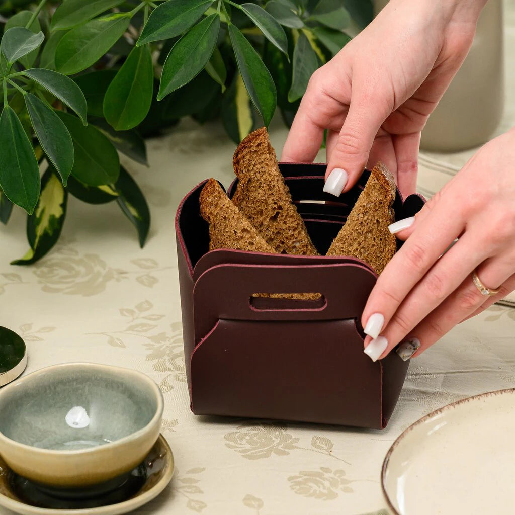 A close-up of a person's hands holding a dark burgundy leather bread basket filled with three slices of toasted rye bread. The basket features a unique handle-like slot design. The scene is set on a beige floral tablecloth with a small bowl and green plant in the background