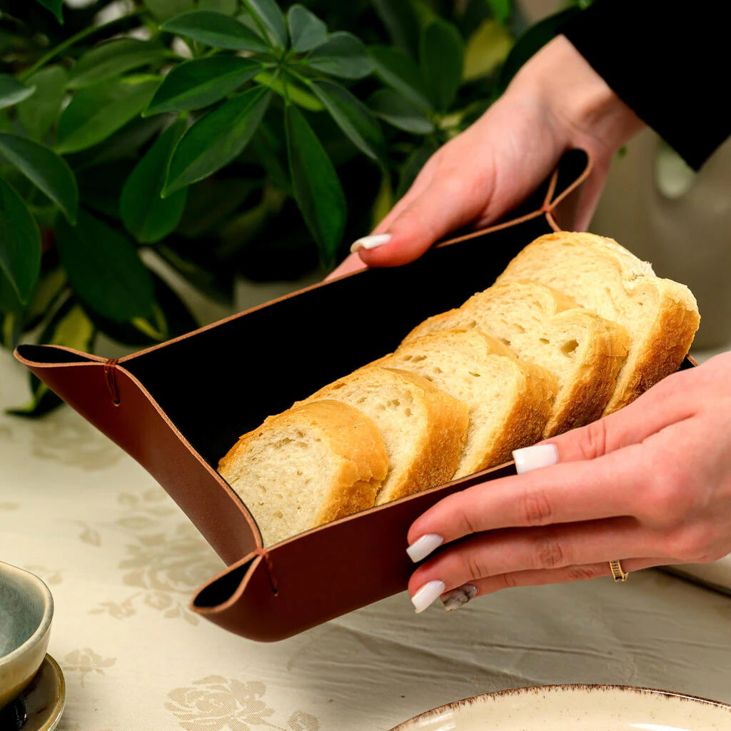 Stylish brown genuine leather bread valet tray on a restaurant table, holding five slices of fresh bread. The image emphasizes the unique folded corner design and the premium feel of the leather, with a hand interacting with the bread for scale and warmth