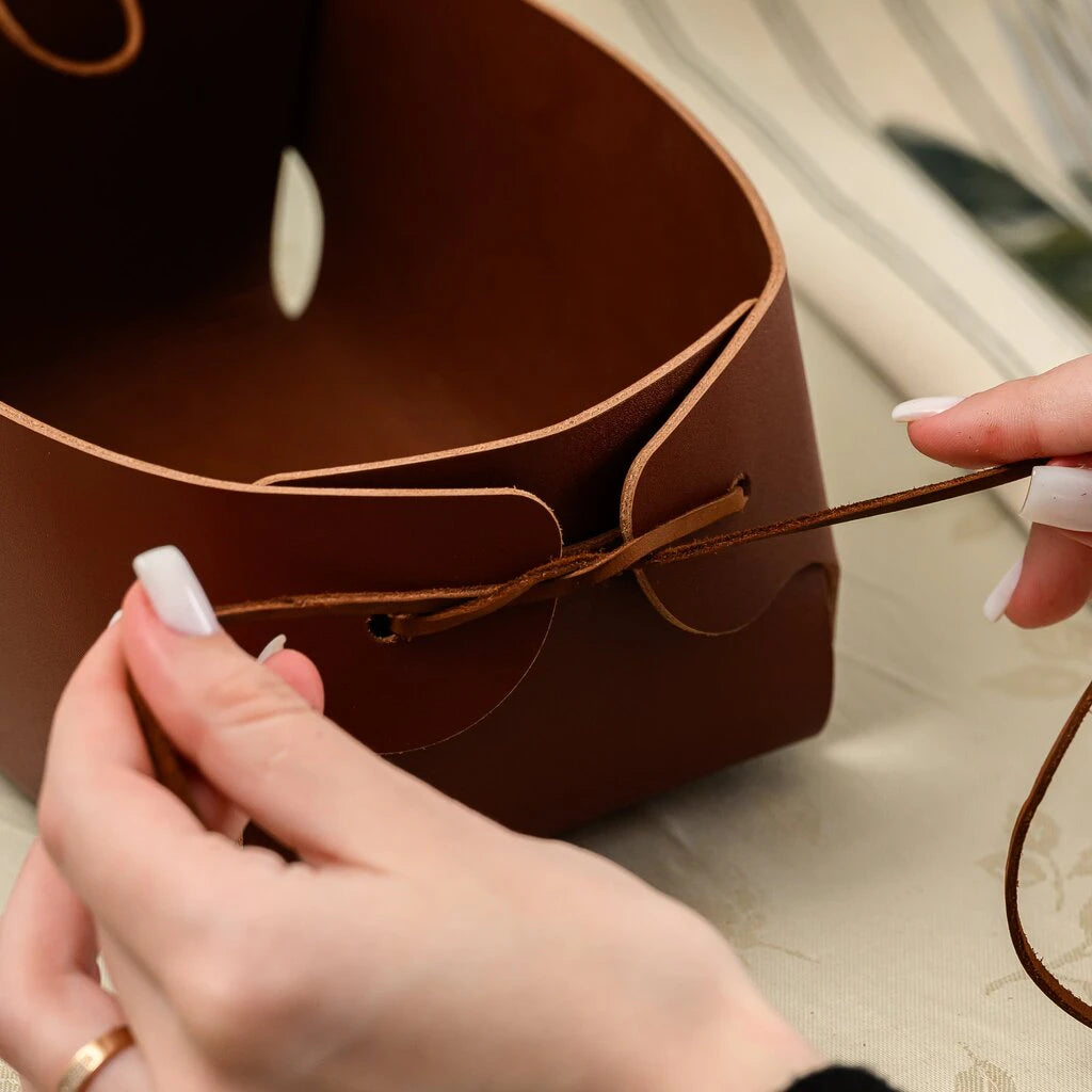 A person assembling a brown leather bread basket by threading a leather cord through side holes