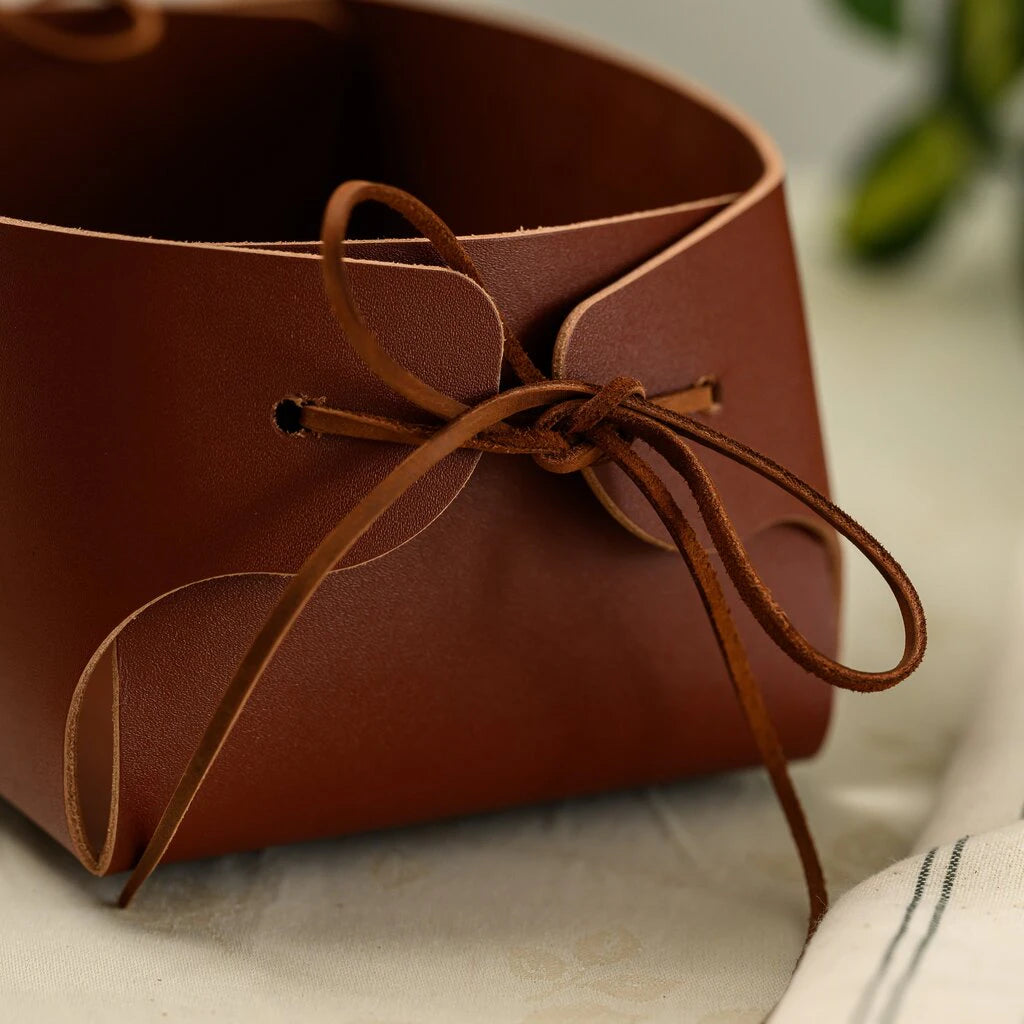 Hands holding a handcrafted brown leather bread basket with elegant side leather ties, showcasing the smooth genuine leather texture and artisanal design