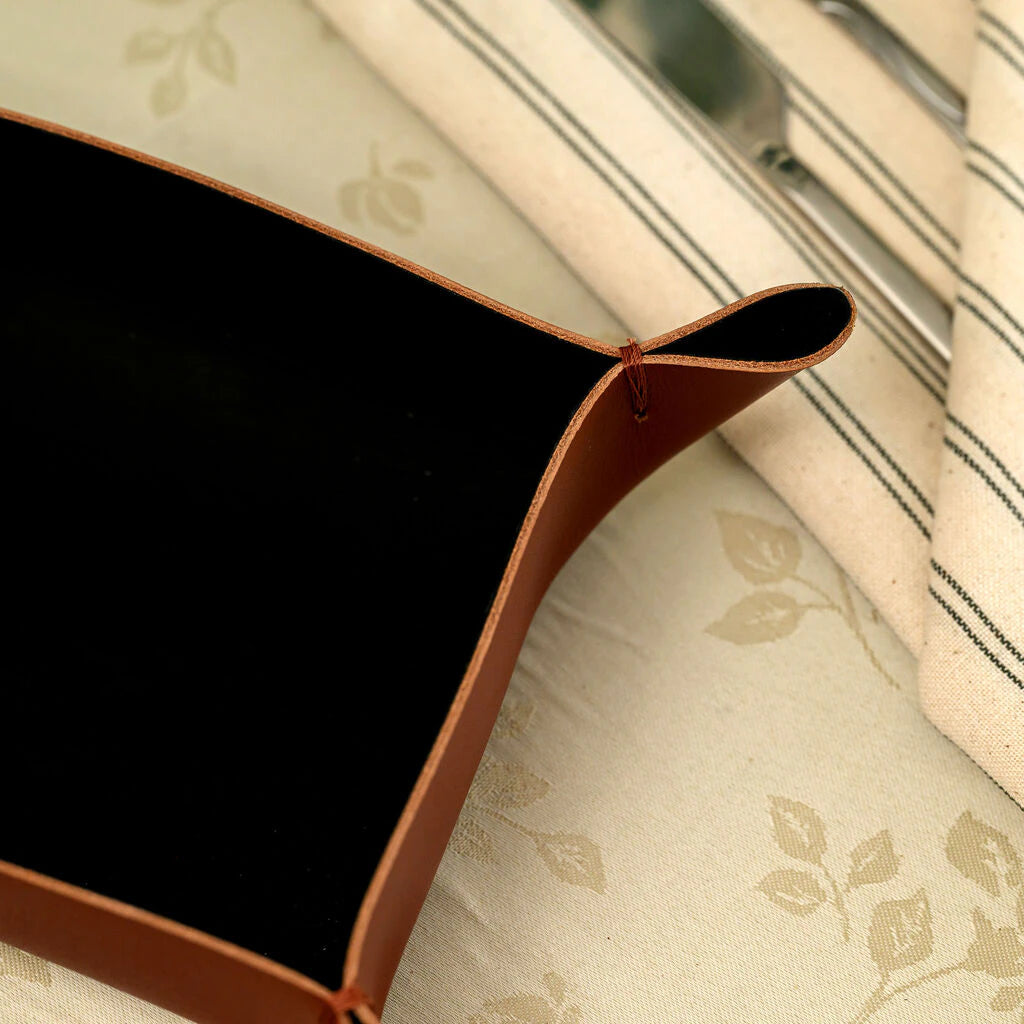 A top-down view of a handcrafted rectangular bread basket made from dark brown genuine leather. The photo highlights its unique construction with folded, stitched corners and a sleek black interior. It is shown on a restaurant table as a premium accessory for serving bread or appetizers in a modern dining setting