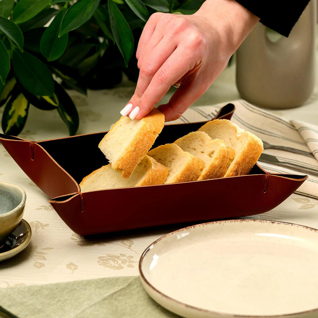 A handcrafted rectangular bread basket made of genuine brown leather with folded, stitched corners is filled with neatly arranged slices of fresh white bread. A woman's hand with a white manicure is gently taking one slice, highlighting the tactile experience and modern rustic aesthetic of the table setting. Perfect for serving bread in upscale restaurants or home dining