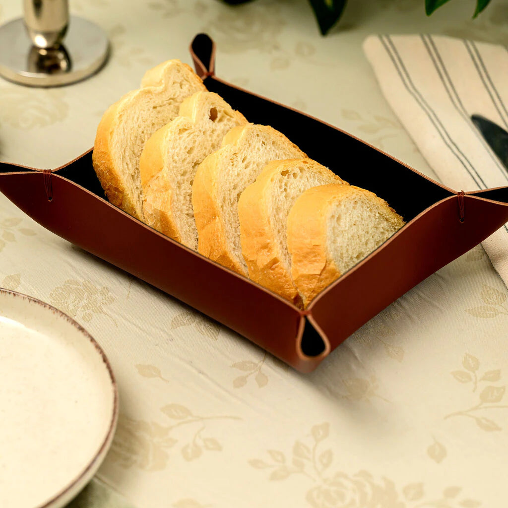 A handcrafted rectangular napkin holder made from high-quality brown genuine leather. The holder features a minimalist open-top design with precise decorative stitching along the edges. It is filled with white paper napkins and placed on a restaurant table, showcasing its elegant and durable construction for professional table setting.