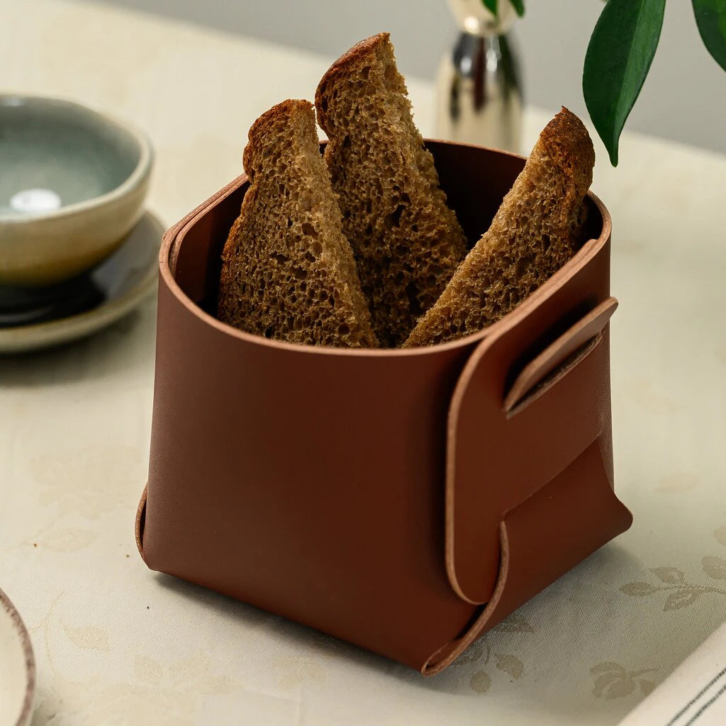 A square-shaped brown leather bread holder containing three thick slices of toasted dark bread. The leather is folded at the corners to create a deep, sturdy container. It sits on a light floral table setting with a small ceramic bowl in the background