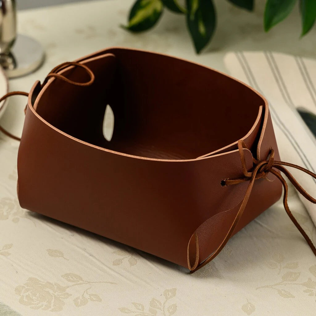 A minimalist handmade bread basket made of smooth brown leather. The sides are folded and secured with thin leather laces tied into knots. It is placed on a light-colored table with a floral pattern and a striped napkin nearby.