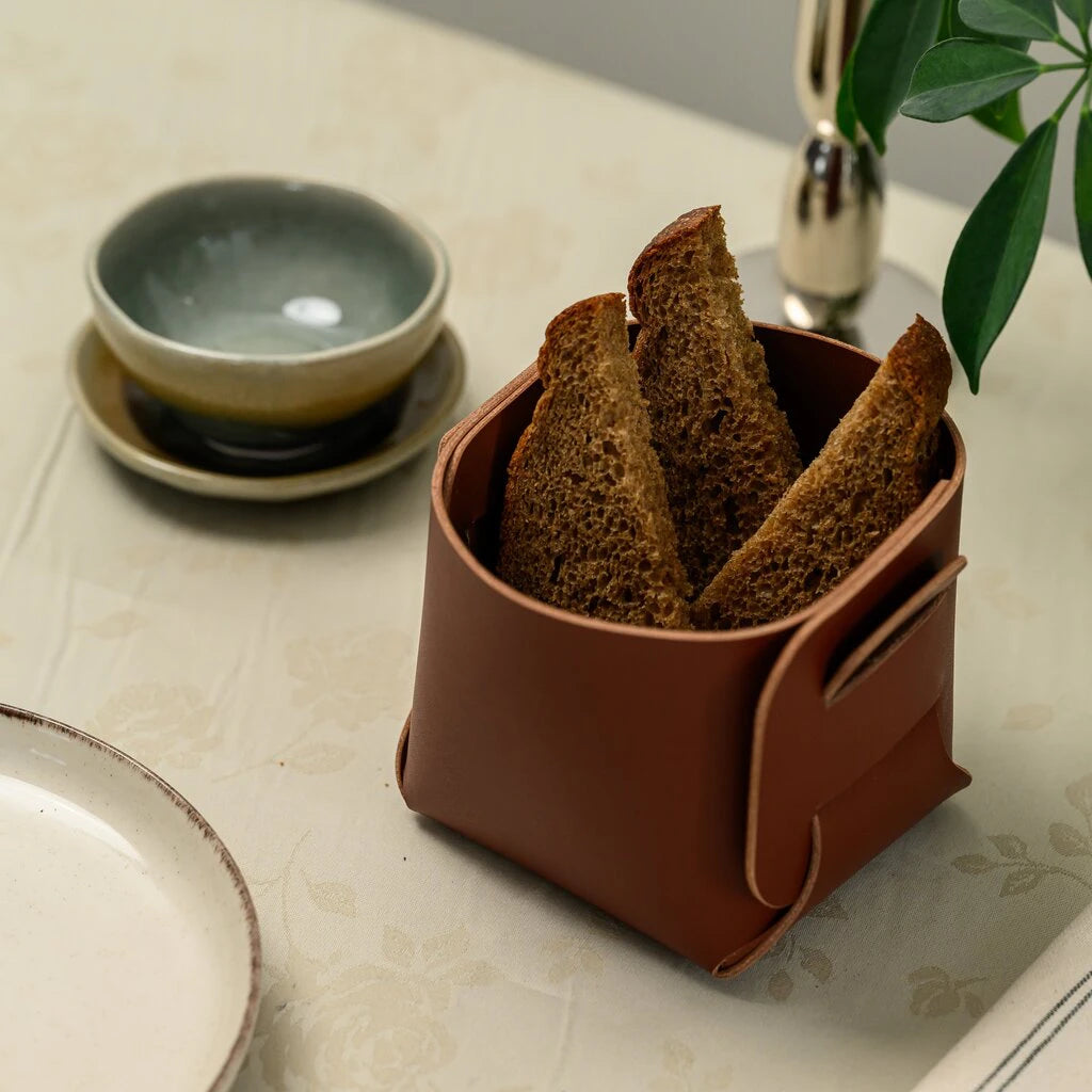 A high-angle shot of a square leather bread bin containing slices of dark rye bread. It is placed on a light-colored tablecloth next to a small olive-green ceramic bowl and a white plate. In the background, a silver candle holder and green leaves are partially visible