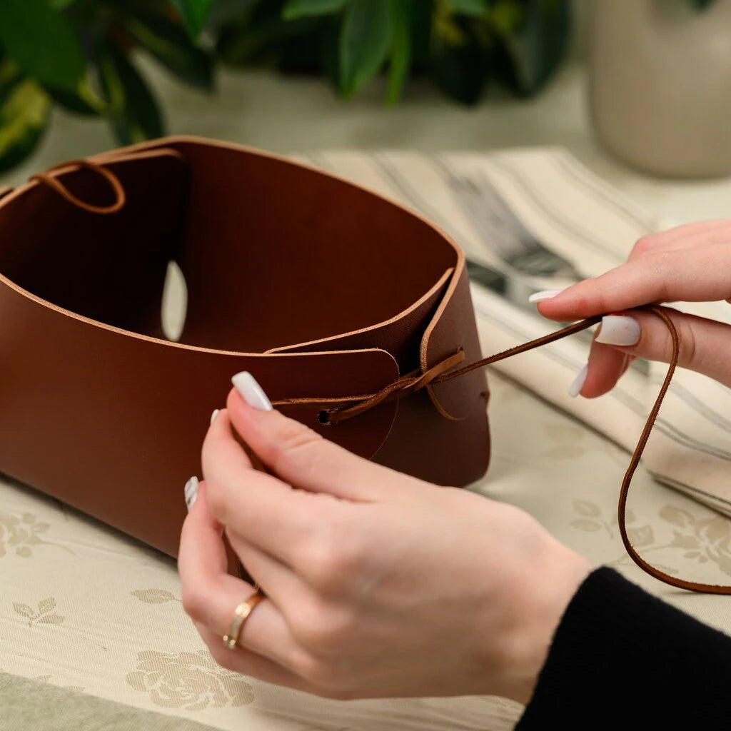 Close-up of a person's hands assembling a minimalist brown leather bread basket. The person is threading a thin leather string through pre-cut holes on the sides to shape the container. The background shows a dining table setting with a linen tablecloth and a plant