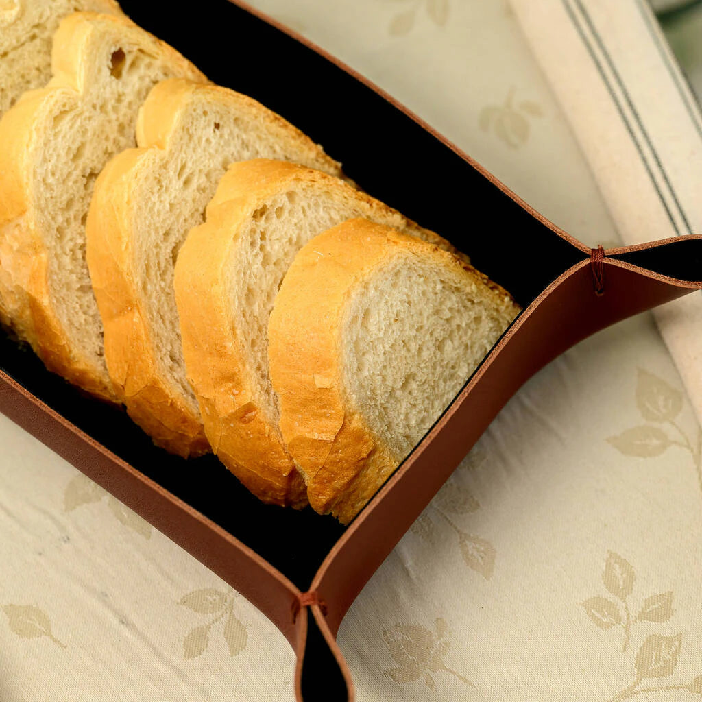 Top view of a handcrafted brown leather bread basket with elegant stitched corners, holding fresh bread slices on a floral tablecloth
