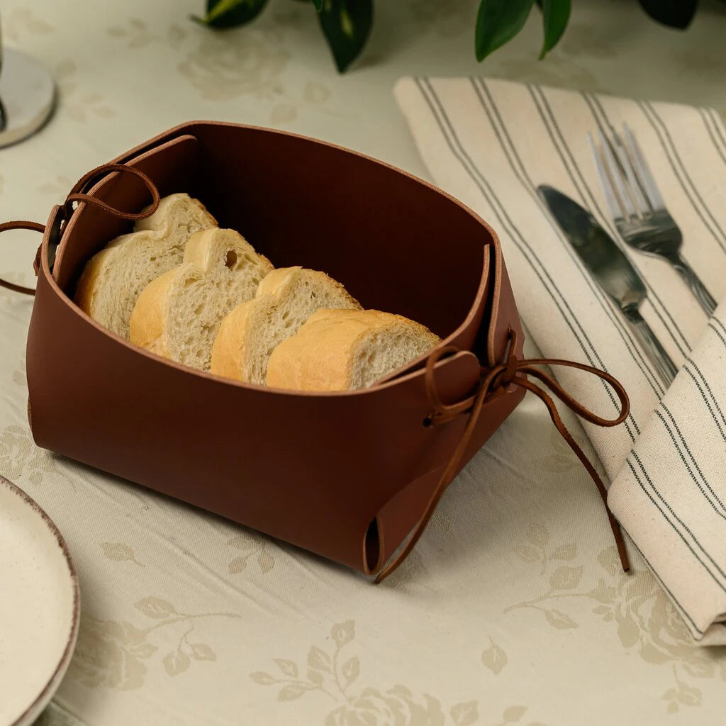 A brown leather bread basket with tied lace corners sitting on a floral tablecloth