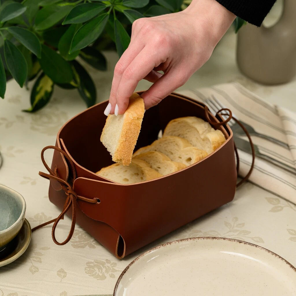 A hand placing a slice of white bread into a brown leather bread basket on a set table