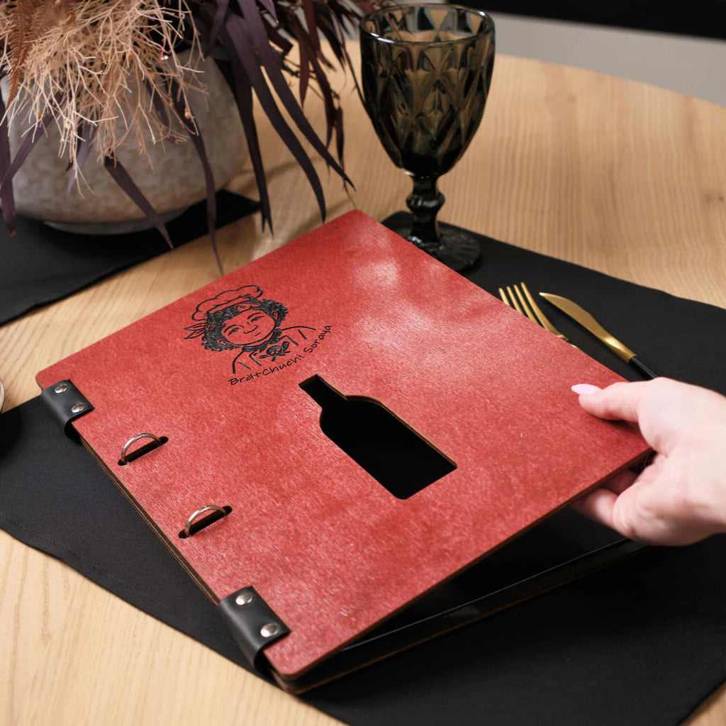 A hand lifting the front cover of a chili-red wooden menu folder, showcasing the black engraved chef logo and the wine bottle-shaped window