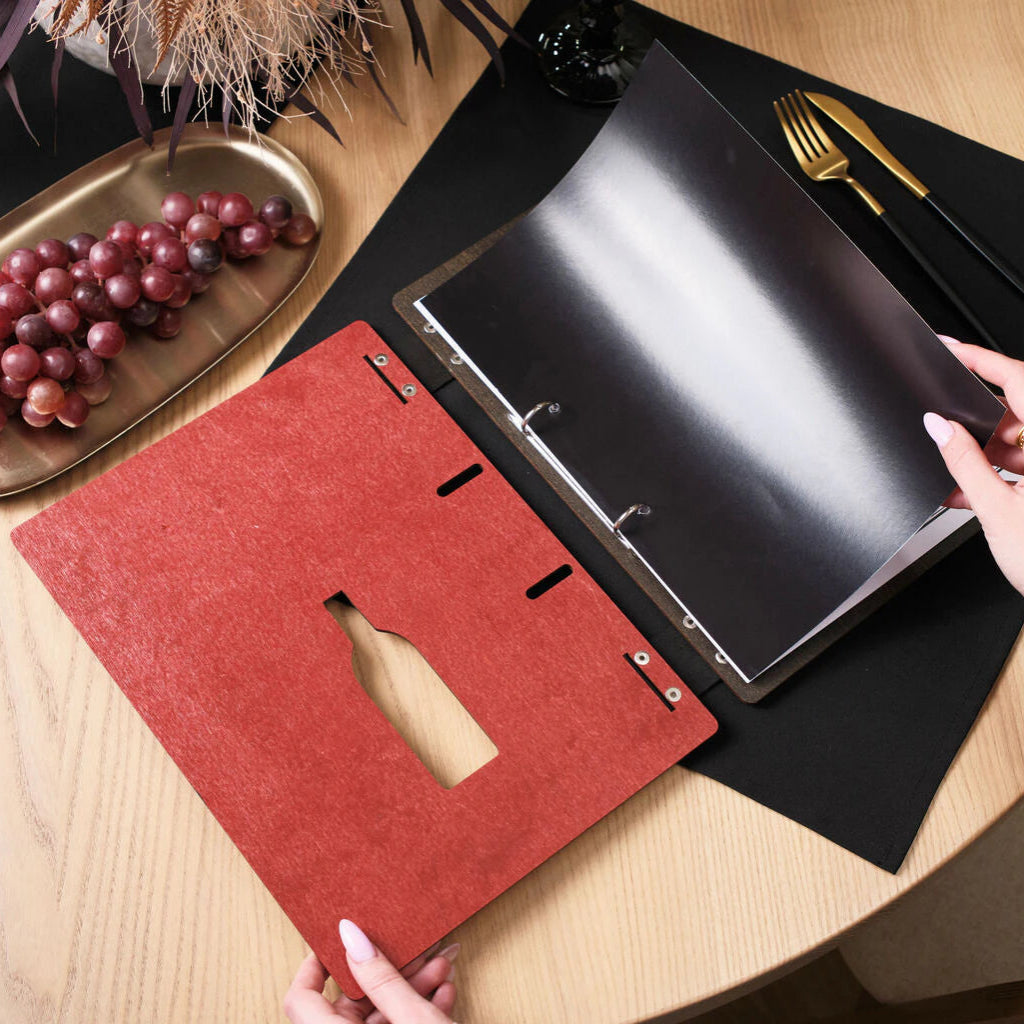 An open view of the chili-red wooden menu folder, showing the inner ring binder holding black menu pages, with a laser-cut wine bottle silhouette visible on the cover.