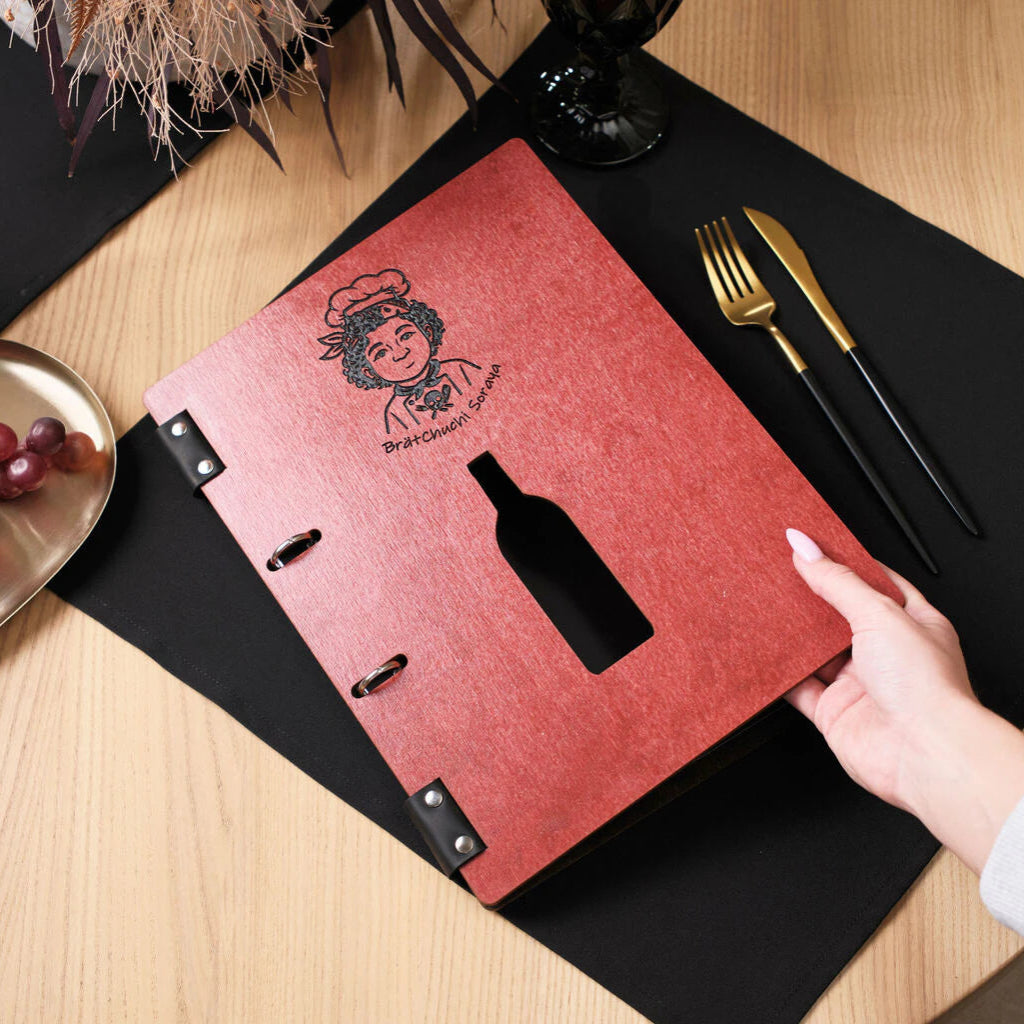 A hand lifting the cover of a chili-red wooden menu folder on a set restaurant table, showcasing the engraved branding and the unique wine bottle-shaped cutout.
