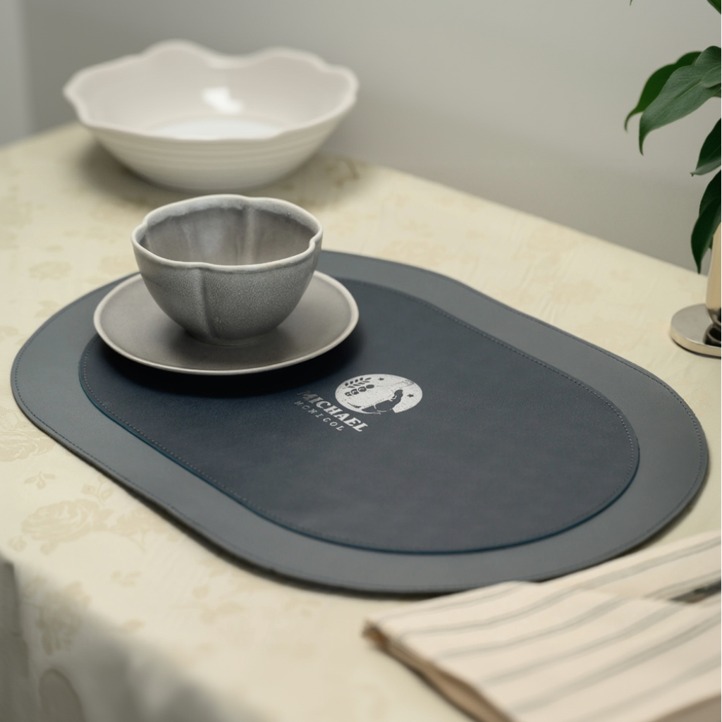 An oval leather placemat with a layered design, featuring a smooth outer rim and a textured inner section joined by fine perimeter stitching. A circular logo with the name "MICHAEL MCNICOL" is centered on the mat. A ceramic bowl on a saucer is placed on the left side of the placemat, which sits on a floral-patterned tablecloth alongside other tableware.