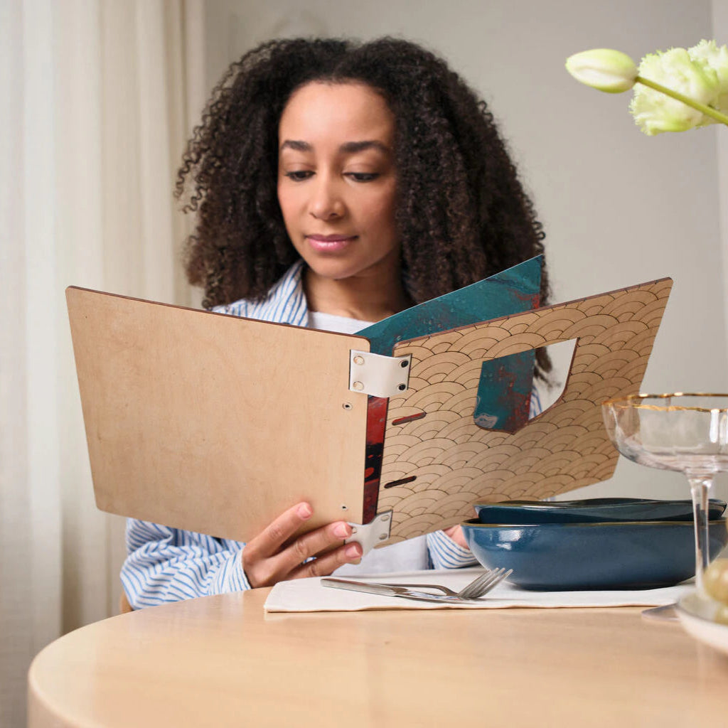 Lifestyle shot of a smiling young woman reading an open handcrafted wooden menu at a restaurant table.