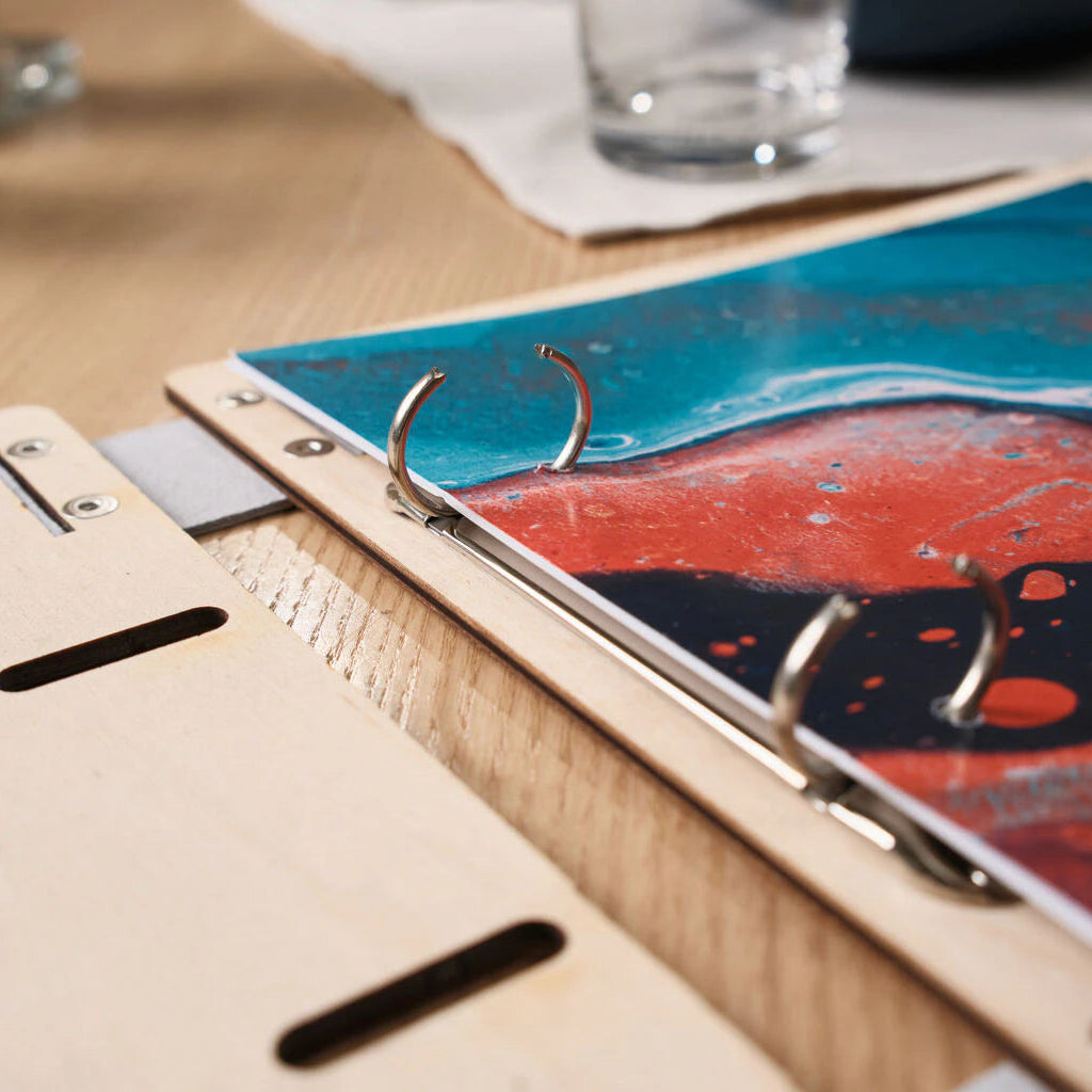 Angled close-up view of the internal ring binder of an open light wood menu holder, highlighting the colorful painted insert and hardware details.