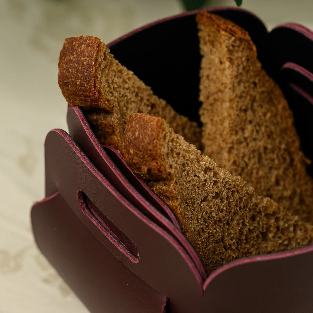 A macro shot of several thick slices of dark rye bread served in a handcrafted burgundy leather basket. The photo focuses on the porous texture of the bread and the smooth, matte finish of the dark red leather edges with purple undertones.