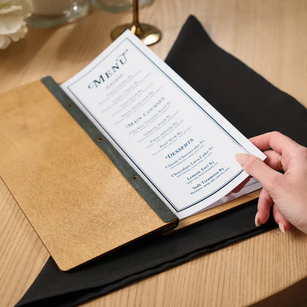 A side-angle view of an open oak-colored wooden menu folder where a person's hand is turning the paper pages, showcasing the slim profile and the $5 fixed pricing for all items.