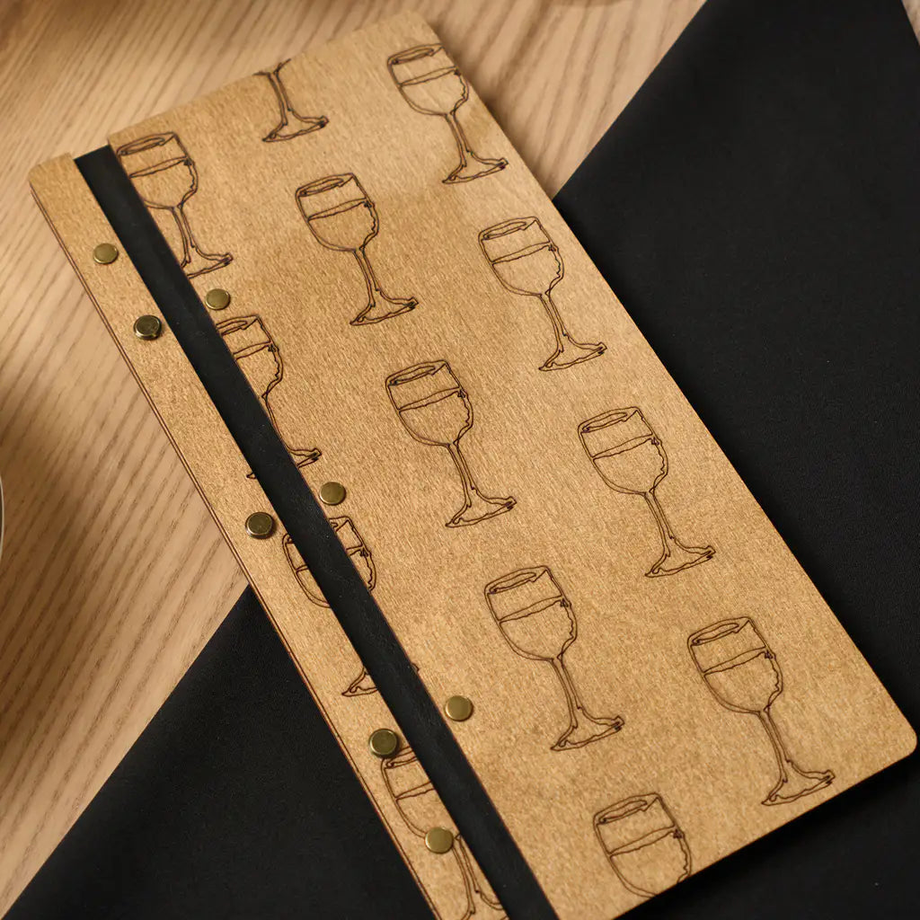 A slim oak-colored wooden menu folder featuring a repetitive engraved pattern of wine glasses. The cover is secured with a black spine and brass-colored rivets, resting on a black napkin