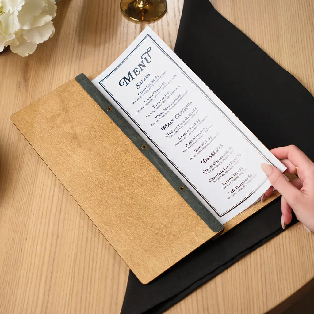 A person’s hand holds the edge of the menu pages within the oak wooden folder, illustrating the tactile quality of the materials and the secure internal binding.