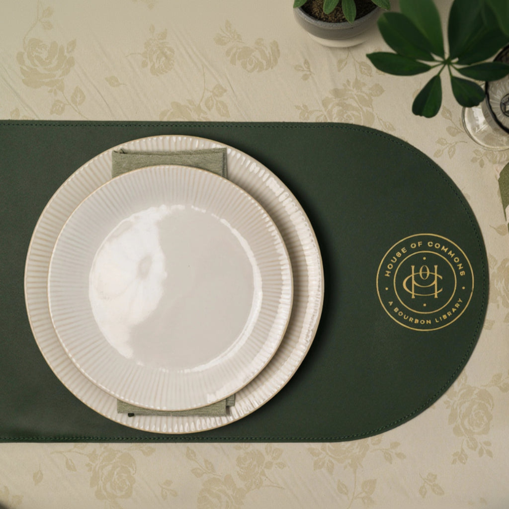 Dark green leather placemat with rounded edges, finished with premium gold foil embossing, paired with white ceramic plates and a linen napkin on a soft floral tablecloth—an elegant and refined restaurant table setting, luxury hospitality decor, perfect for fine dining, cafes, weddings, and upscale hospitality interiors