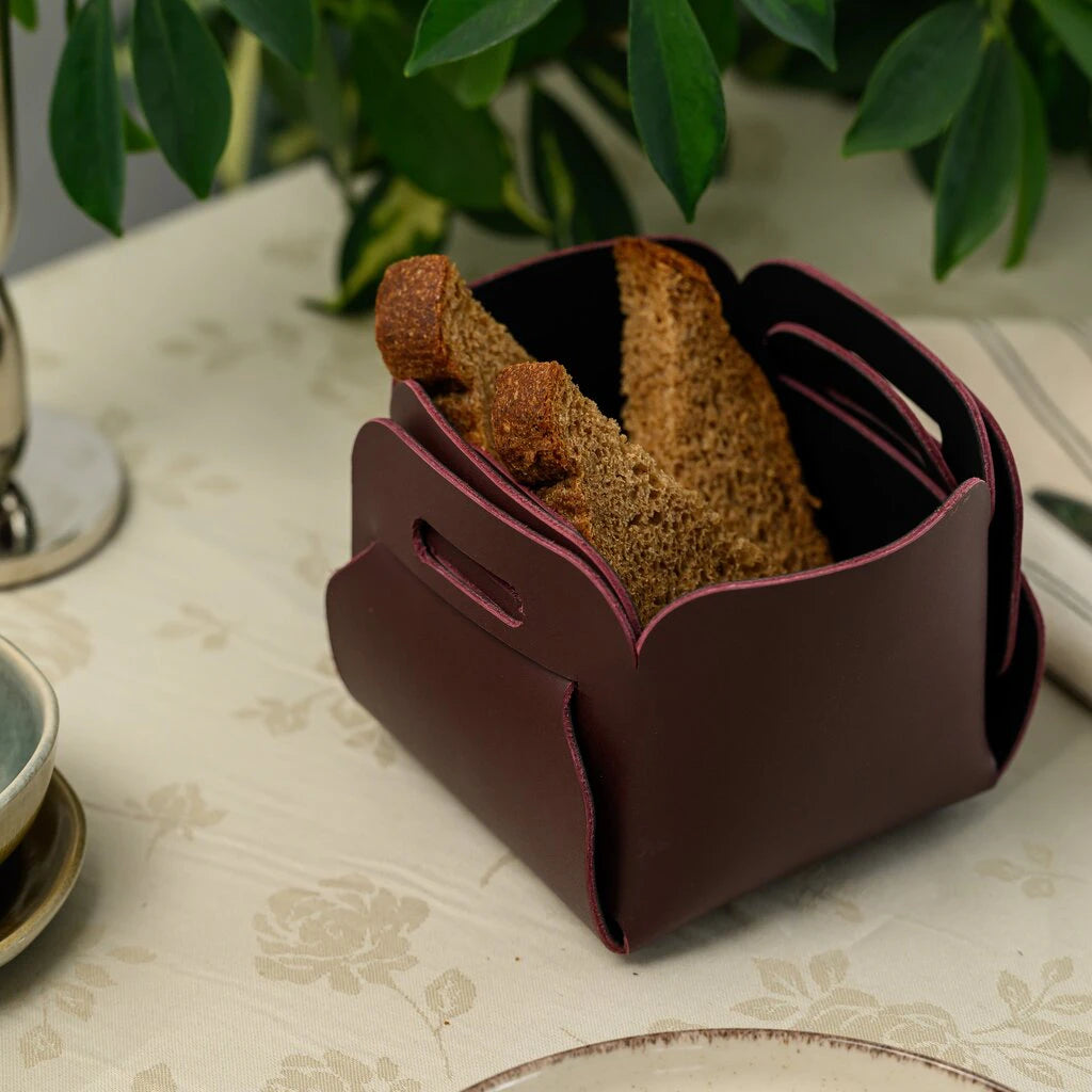 A deep burgundy leather bread basket filled with slices of dark rye bread, featuring a unique interlocking design without stitching